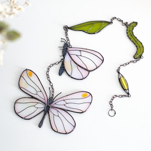 May include: Stained glass butterfly suncatcher with white and pink wings, yellow accents, and black outlines. The butterfly is connected to a chain with green leaves and a yellow teardrop shape. Decorative home decor.