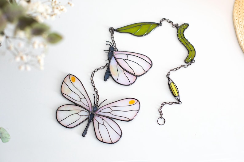 May include: Stained glass butterfly suncatcher with white and pink wings, yellow accents, and black outlines. The butterfly is connected to a chain with green leaves and a yellow teardrop shape. Decorative home decor.
