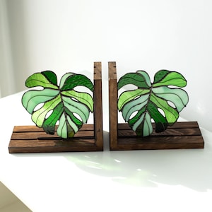 K&ouml;nnte beinhalten: Zwei Buchst&uuml;tzen aus Holz mit Monstera-Bl&auml;ttern aus Buntglas in Gr&uuml;nt&ouml;nen. Die Bl&auml;tter sind auf einer dunkelbraunen, rechteckigen Basis angeordnet. Die Buchst&uuml;tzen dienen dazu, B&uuml;cher aufrecht zu halten.