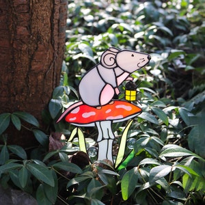 May include: A stained glass mouse holding a gift sits on a red and white mushroom. The mushroom is surrounded by green leaves.