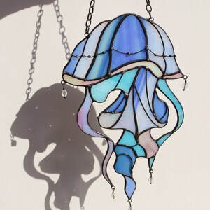 Op de afbeelding: Een gebrandschilderd glas-zonnevanger in de vorm van een kwal met een blauw, paars en roze kleurenschema. De kwal hangt aan een zwarte ketting en heeft kristallen kralen die eronder hangen.