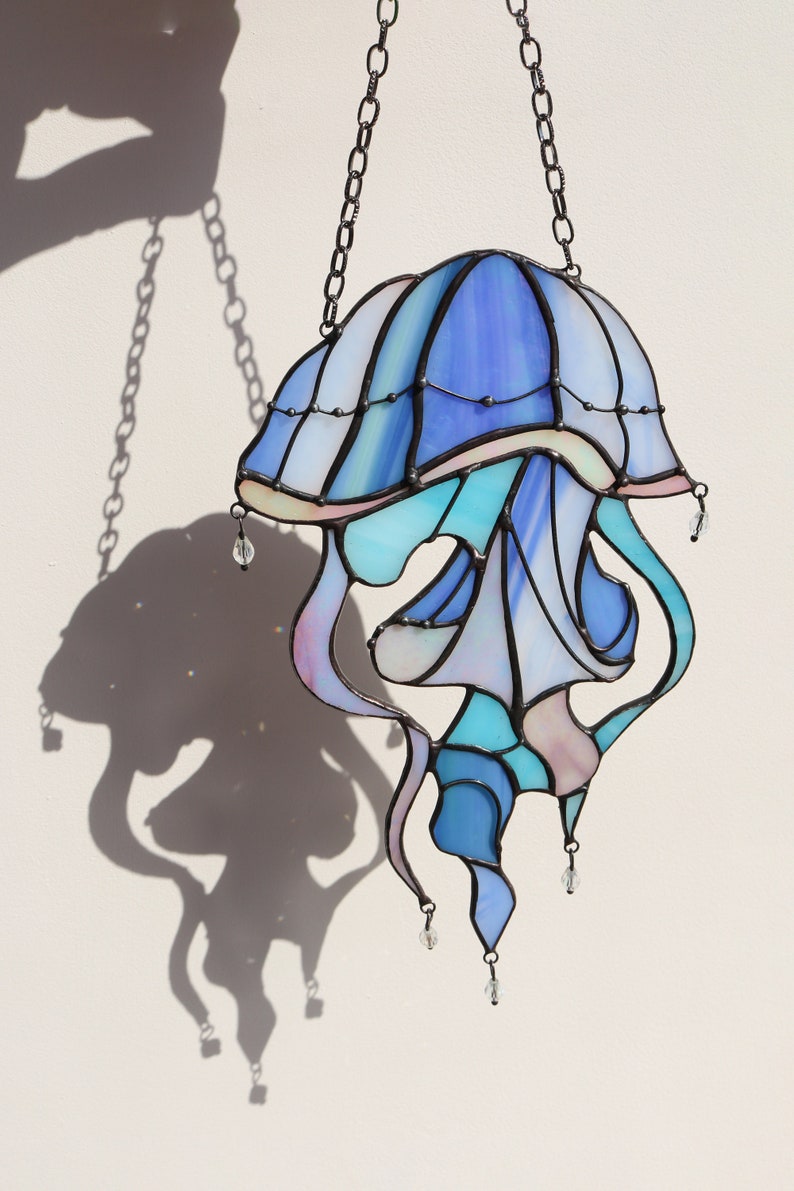 Op de afbeelding: Een gebrandschilderd glas-zonnevanger in de vorm van een kwal met een blauw, paars en roze kleurenschema. De kwal hangt aan een zwarte ketting en heeft kristallen kralen die eronder hangen.