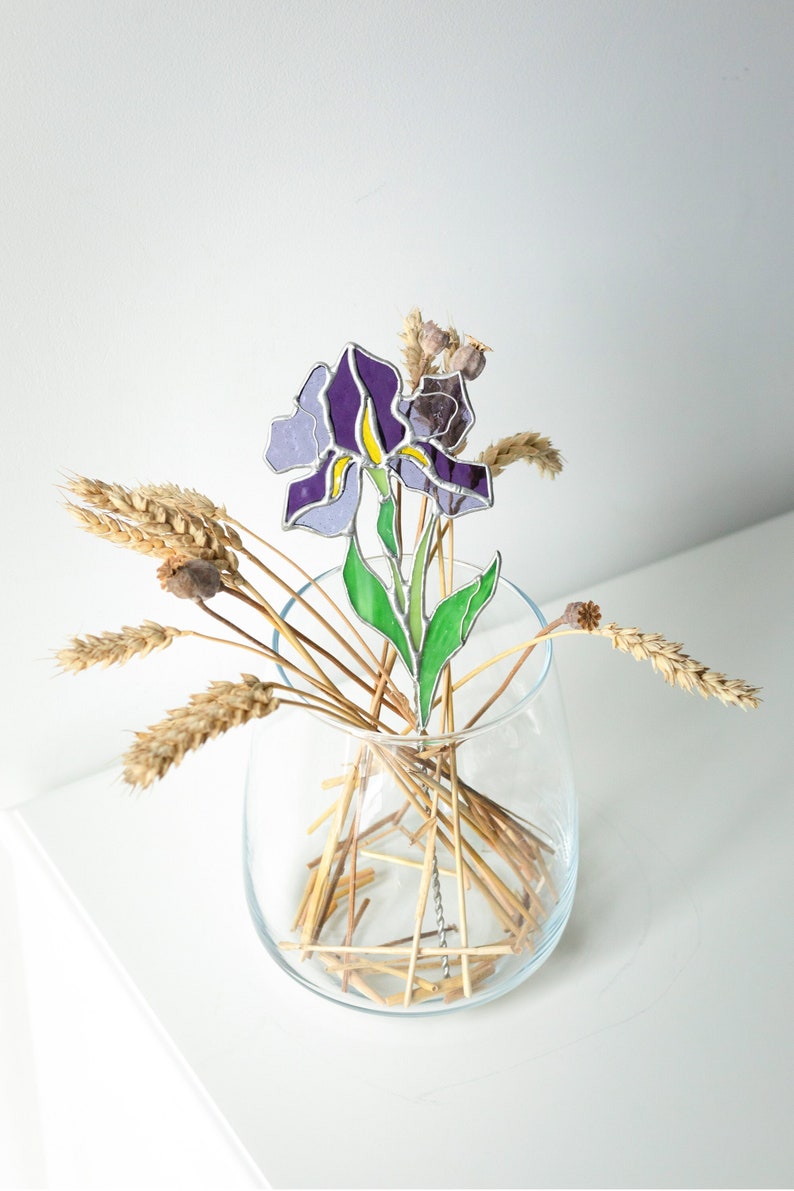 May include: A stained glass iris flower in purple and green, in a clear glass vase with dried wheat stalks.