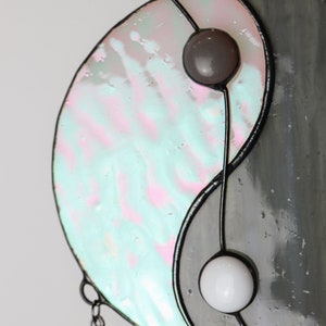 May include: A stained glass yin yang suncatcher with a gray and iridescent blue and pink design. The suncatcher has two small glass beads in the center.