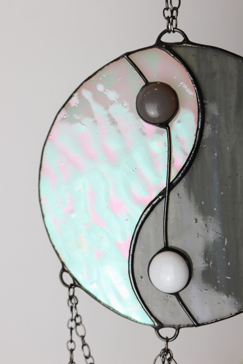 May include: A stained glass yin yang suncatcher with a gray and iridescent blue and pink design. The suncatcher has two small glass beads in the center.