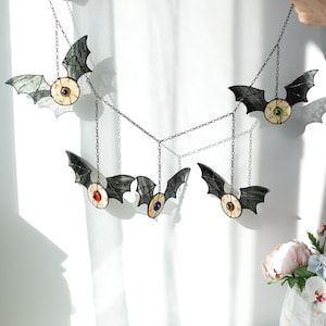 May include: A hanging mobile featuring five stained glass bats with outstretched wings. Each bat has a central circular eye with a distinct iris colour, including red, blue, green, and brown, all connected by thin dark metal chains for display.