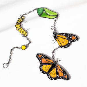 May include: A decorative chain featuring a yellow caterpillar, a green chrysalis, and two orange and black monarch butterflies. The chain is made of a dark metal and is set against a white background.