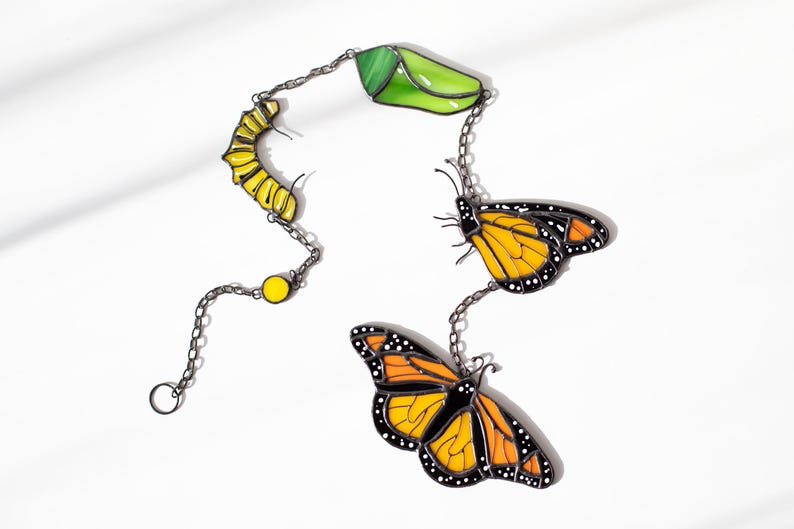 May include: A decorative chain featuring a yellow caterpillar, a green chrysalis, and two orange and black monarch butterflies. The chain is made of a dark metal and is set against a white background.