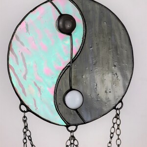 May include: A stained glass yin yang suncatcher with iridescent blue and pink glass on one side and gray glass on the other side. The suncatcher is hanging from a black chain.