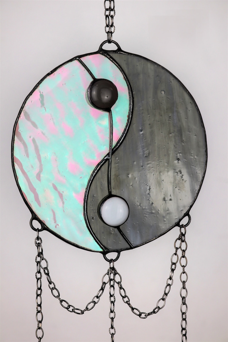 May include: A stained glass yin yang suncatcher with iridescent blue and pink glass on one side and gray glass on the other side. The suncatcher is hanging from a black chain.