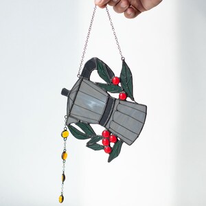 Puede incluir: Un atrapasue&ntilde;os de vidrieras con forma de cafetera italiana, con paneles de vidrio gris, hojas de color verde oscuro y bayas de color rojo brillante. Una fina cadena de metal cuelga de la parte inferior con peque&ntilde;as cuentas de vidrio &aacute;mbar.
