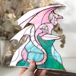Op de afbeelding: Gebrandschilderde glazen drakenbeeld in roze, wit en blauwgroen. De draak zit op een kleurrijke basis van groen en blauw glas. De vleugels zijn gespreid en de draak is gedetailleerd met zilveren soldeer.