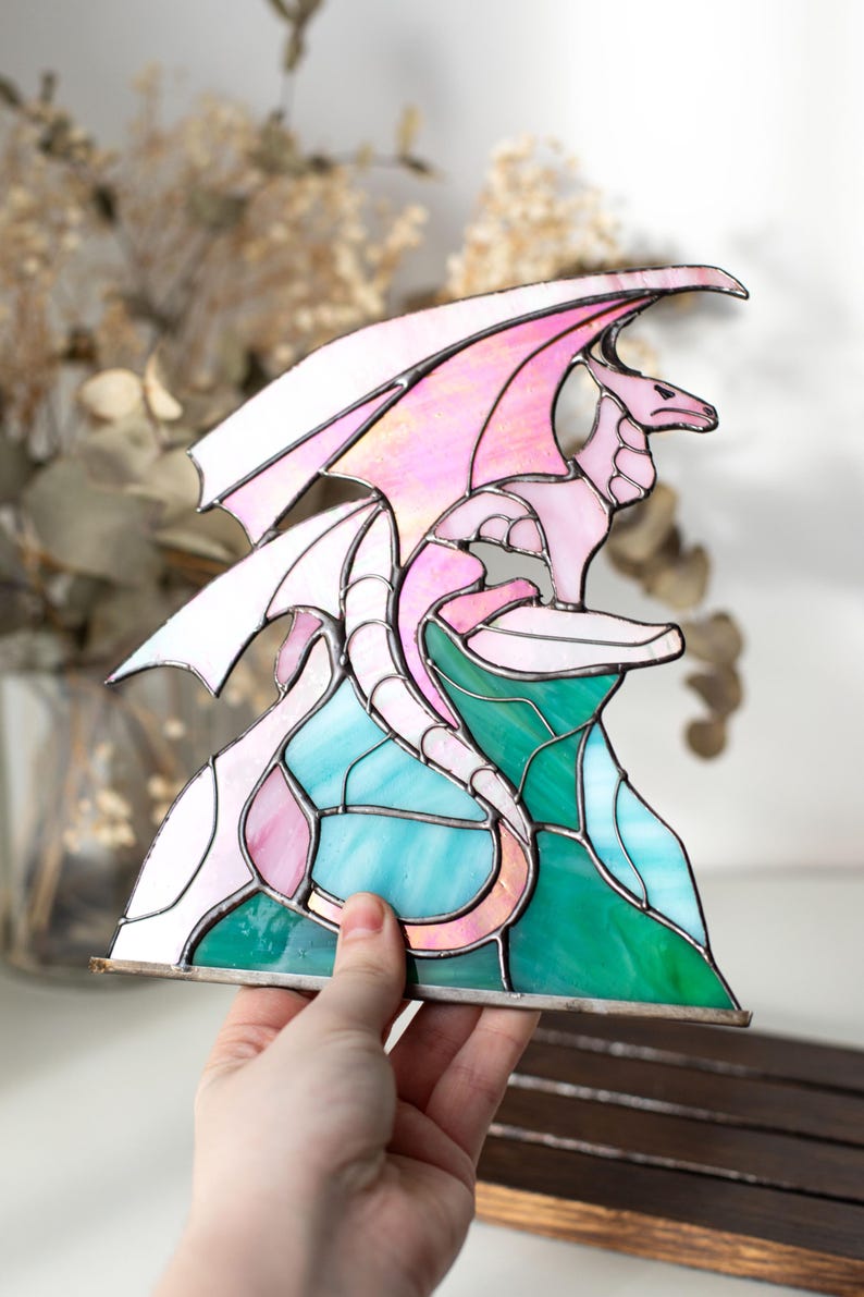 Op de afbeelding: Gebrandschilderde glazen drakenbeeld in roze, wit en blauwgroen. De draak zit op een kleurrijke basis van groen en blauw glas. De vleugels zijn gespreid en de draak is gedetailleerd met zilveren soldeer.
