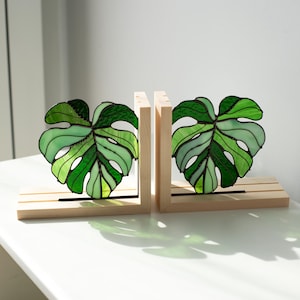 K&ouml;nnte beinhalten: Zwei Buchst&uuml;tzen aus Holz mit Glasmalerei-Monstera-Bl&auml;ttern in Gr&uuml;nt&ouml;nen. Die Bl&auml;tter sind in einem Holzrahmen mit horizontaler Basis angeordnet. Die Buchst&uuml;tzen sind zum Aufstellen von B&uuml;chern gedacht.