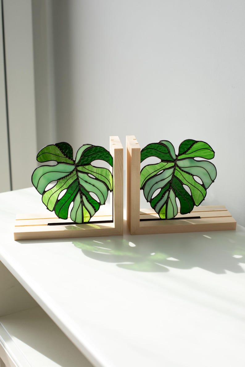 K&ouml;nnte beinhalten: Zwei Buchst&uuml;tzen aus Holz mit Glasmalerei-Monstera-Bl&auml;ttern in Gr&uuml;nt&ouml;nen. Die Bl&auml;tter sind in einem Holzrahmen mit horizontaler Basis angeordnet. Die Buchst&uuml;tzen sind zum Aufstellen von B&uuml;chern gedacht.