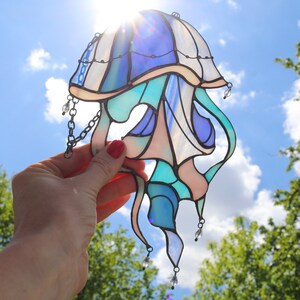 Op de afbeelding: Een gebrandschilderd glas-zonnevanger in de vorm van een kwal met blauwe, groene en roze glasstukken. De zonnevanger heeft een ketting en kristallen kralen die eraan hangen.
