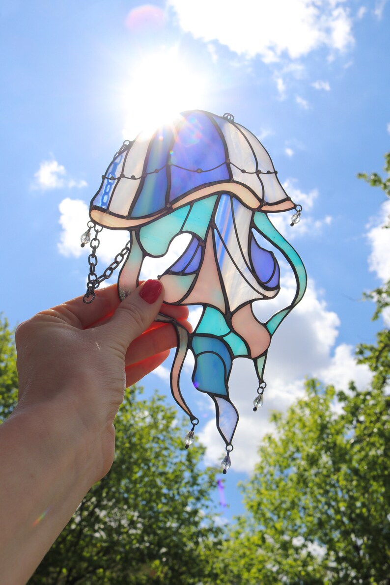 Op de afbeelding: Een gebrandschilderd glas-zonnevanger in de vorm van een kwal met blauwe, groene en roze glasstukken. De zonnevanger heeft een ketting en kristallen kralen die eraan hangen.
