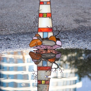 May include: A stained glass lighthouse with red, orange, and white stripes, reflected in a puddle of water. The lighthouse is surrounded by brown, orange, and pink stained glass rocks.