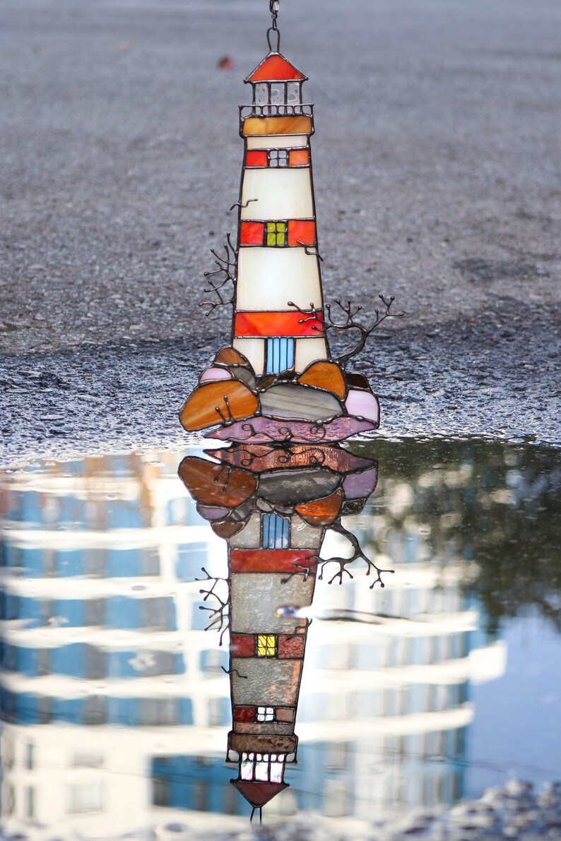 May include: A stained glass lighthouse with red, orange, and white stripes, reflected in a puddle of water. The lighthouse is surrounded by brown, orange, and pink stained glass rocks.