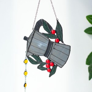 Puede incluir: Un atrapasoles de vidrieras con forma de cafetera moka en tonos grises, acentuado con hojas verdes y bayas rojas brillantes. Una delicada cadena de metal cuelga del pico con peque&ntilde;as cuentas de vidrio amarillas y &aacute;mbar adjuntas.