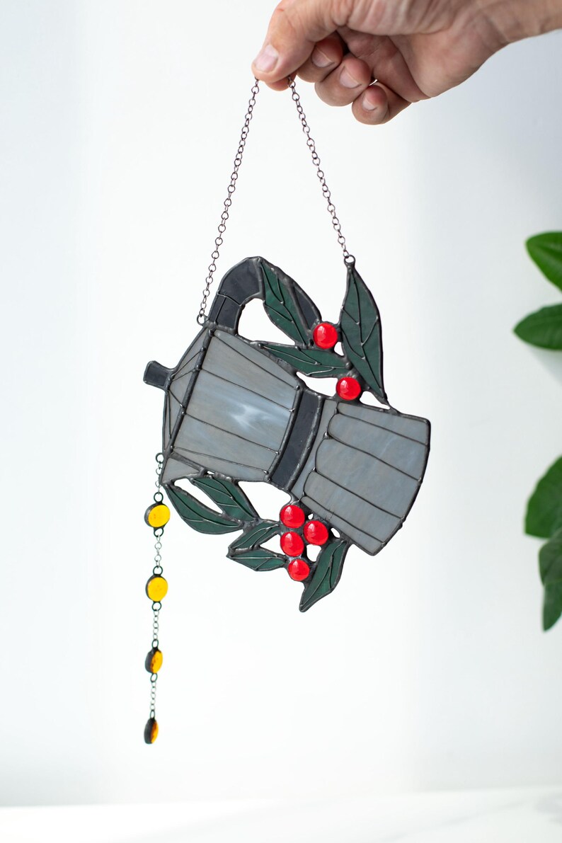 Puede incluir: Un atrapasoles de vidrieras con forma de cafetera moka en tonos grises, acentuado con hojas verdes y bayas rojas brillantes. Una delicada cadena de metal cuelga del pico con peque&ntilde;as cuentas de vidrio amarillas y &aacute;mbar adjuntas.