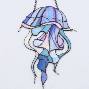 Op de afbeelding: Een gebrandschilderd glas-zonnevanger in de vorm van een kwal met iriserende blauwe, paarse en roze glazen. De kwal heeft een ketting om op te hangen en is een decoratief stuk voor een raam of muur.
