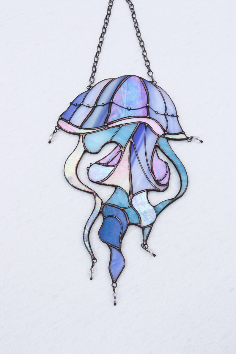 Op de afbeelding: Een gebrandschilderd glas-zonnevanger in de vorm van een kwal met iriserende blauwe, paarse en roze glazen. De kwal heeft een ketting om op te hangen en is een decoratief stuk voor een raam of muur.