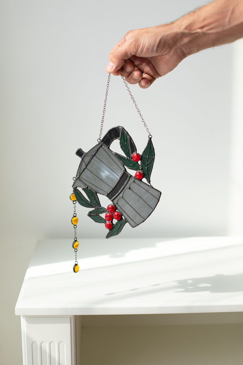 Puede incluir: Un atrapasue&ntilde;os de vidrieras con forma de cafetera moka, con paneles de vidrio gris, hojas de color verde oscuro y bayas de color rojo brillante. Una cadena vertical de peque&ntilde;as cuentas de vidrio amarillas cuelga del pico, suspendida por una cadena de metal.