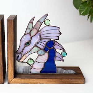May include: Stained glass dragon bookends. The dragon features a lavender head, blue accents, and green glass accents. The bookends are set in a wooden frame. The dragon's head is on the left bookend, and the tail is on the right.