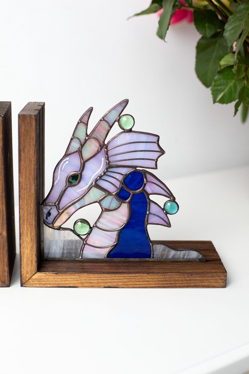 May include: Stained glass dragon bookends. The dragon features a lavender head, blue accents, and green glass accents. The bookends are set in a wooden frame. The dragon's head is on the left bookend, and the tail is on the right.