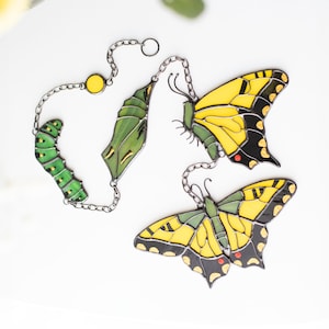May include: Decorative butterfly-themed ornaments. The set includes a caterpillar, chrysalis, and two butterflies. The butterflies have yellow and black wings. The ornaments are connected by silver chains and are set against a white background.