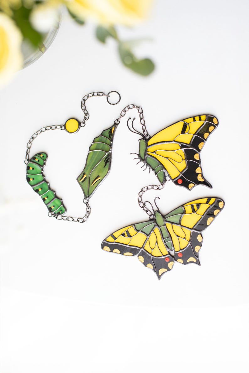 May include: Decorative butterfly-themed ornaments. The set includes a caterpillar, chrysalis, and two butterflies. The butterflies have yellow and black wings. The ornaments are connected by silver chains and are set against a white background.