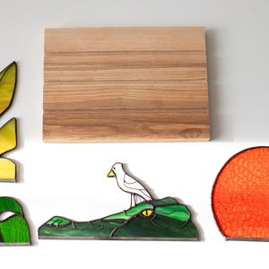 Op de afbeelding: Een assortiment glas-in-lood kunstwerken, waaronder een blad, een paar groene bladeren, een vogel op een alligator en een rode zon. Er is ook een houten snijplank aanwezig. De stukken staan tegen een witte achtergrond.