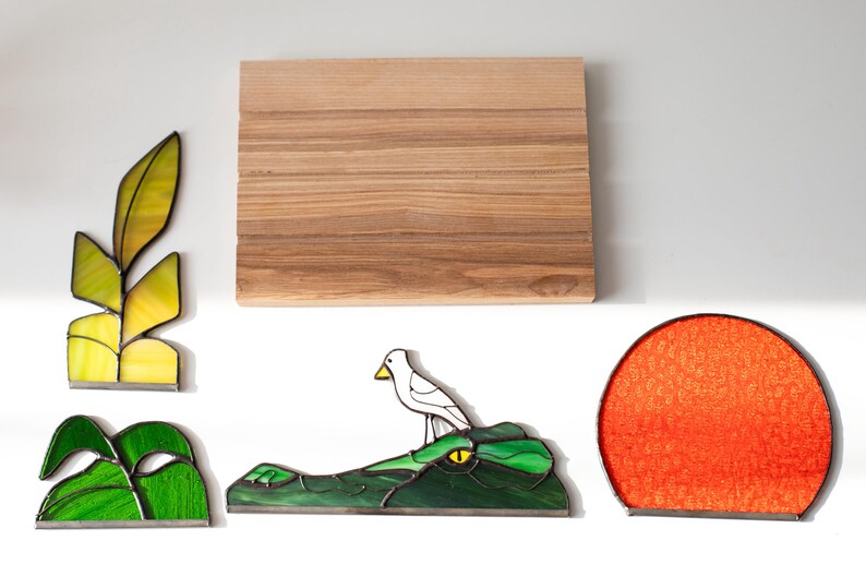 Op de afbeelding: Een assortiment glas-in-lood kunstwerken, waaronder een blad, een paar groene bladeren, een vogel op een alligator en een rode zon. Er is ook een houten snijplank aanwezig. De stukken staan tegen een witte achtergrond.