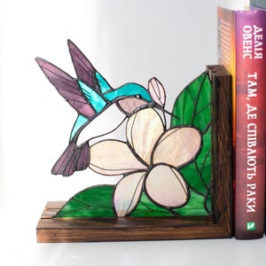 Pu&ograve; includere: Reggilibri in vetro colorato con un colibr&igrave; e un fiore. Il colibr&igrave; ha dettagli turchese, viola e bianchi. Il fiore &egrave; bianco e rosa, con foglie verdi. I reggilibri sono in legno e sostengono libri.