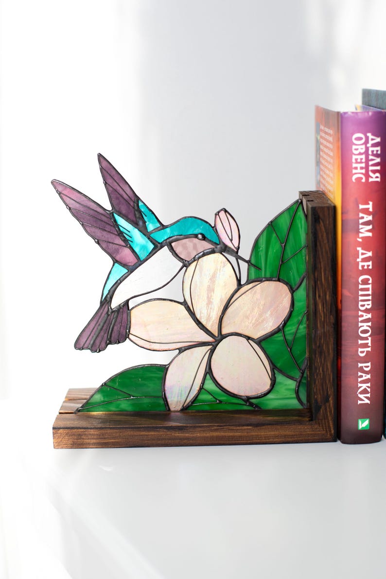 Pu&ograve; includere: Reggilibri in vetro colorato con un colibr&igrave; e un fiore. Il colibr&igrave; ha dettagli turchese, viola e bianchi. Il fiore &egrave; bianco e rosa, con foglie verdi. I reggilibri sono in legno e sostengono libri.