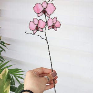 K&ouml;nnte beinhalten: Eine rosa gef&auml;rbte Glasorchidee mit einer wei&szlig;en Perle an der Spitze des Stiels. Die Blume ist an einem langen, gedrehten Draht befestigt.