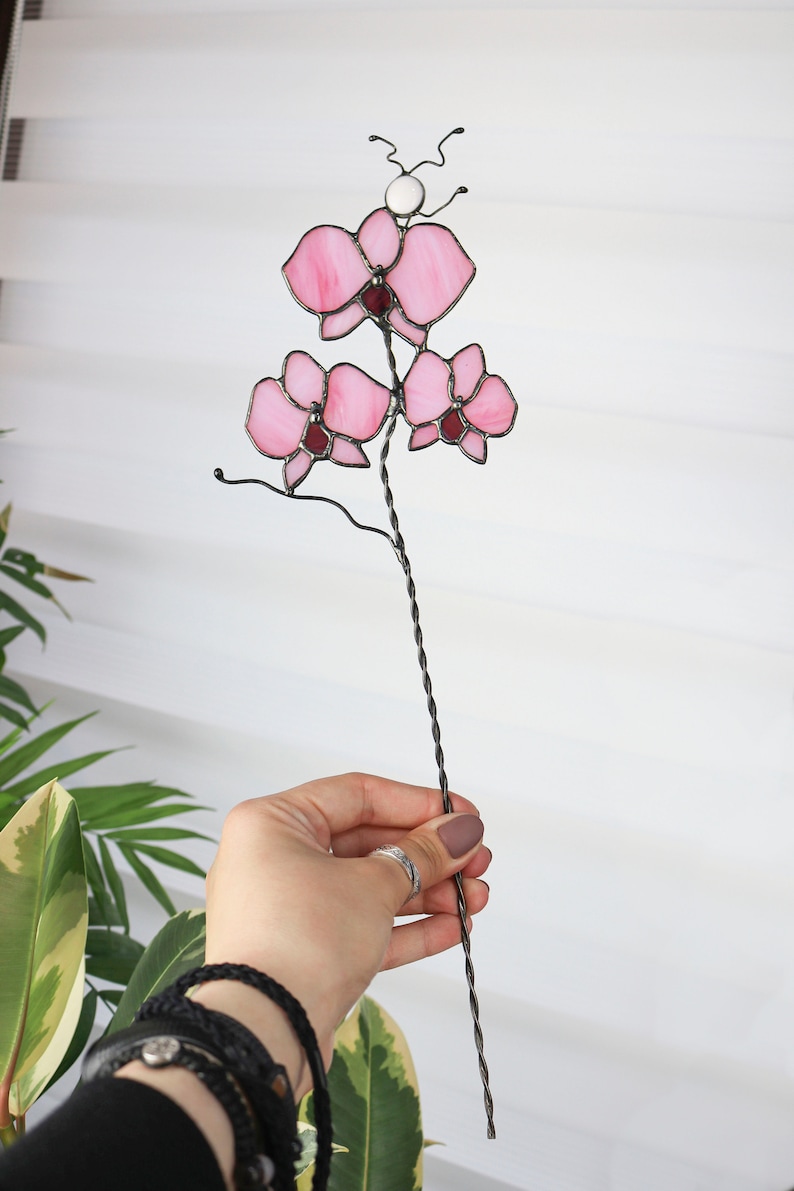 K&ouml;nnte beinhalten: Eine rosa gef&auml;rbte Glasorchidee mit einer wei&szlig;en Perle an der Spitze des Stiels. Die Blume ist an einem langen, gedrehten Draht befestigt.
