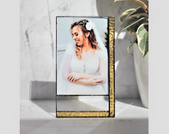 Könnte beinhalten: Ein goldfarbener Bilderrahmen aus Glas mit einem Foto einer Frau in einem weißen Kleid und Schleier. Der Rahmen steht auf einer weißen Oberfläche.
