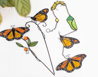 Puede incluir: Arte decorativo de metal con mariposas y orugas. Las mariposas tienen alas naranjas y negras con puntos blancos. La oruga es amarilla y verde. El arte está sobre un fondo blanco.