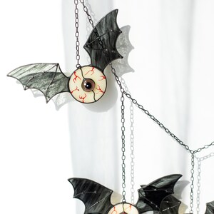 May include: A hanging mobile featuring three stained glass bat figures with realistic eyeball centres. Each bat has dark, translucent wings and a round eye with red veins. The eyes feature brown, orange, and blue irises, all suspended by metal chains.