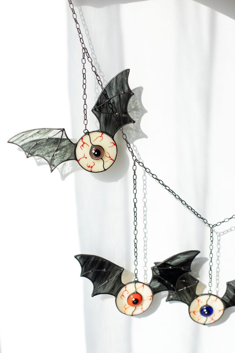 May include: A hanging mobile featuring three stained glass bat figures with realistic eyeball centres. Each bat has dark, translucent wings and a round eye with red veins. The eyes feature brown, orange, and blue irises, all suspended by metal chains.