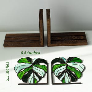 K&ouml;nnte beinhalten: Zwei Buchst&uuml;tzen aus gebeiztem Holz und zwei dekorative Buchst&uuml;tzen aus Glasmalerei in Form von Monstera-Bl&auml;ttern. Die Monstera-Blatt-Buchst&uuml;tzen sind 14 cm hoch und weisen Gr&uuml;n- und Wei&szlig;t&ouml;ne auf. Die Holz-Buchst&uuml;tzen sind dunkelbraun.