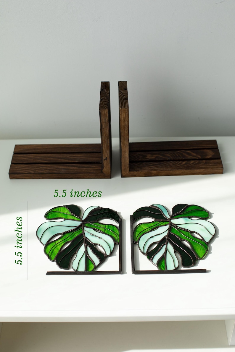 K&ouml;nnte beinhalten: Zwei Buchst&uuml;tzen aus gebeiztem Holz und zwei dekorative Buchst&uuml;tzen aus Glasmalerei in Form von Monstera-Bl&auml;ttern. Die Monstera-Blatt-Buchst&uuml;tzen sind 14 cm hoch und weisen Gr&uuml;n- und Wei&szlig;t&ouml;ne auf. Die Holz-Buchst&uuml;tzen sind dunkelbraun.