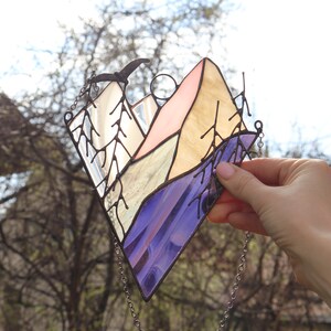 May include: A stained glass window hanging with a chain. The window depicts a mountain range with a pink, yellow, and blue color scheme. A small bird is flying above the mountains.