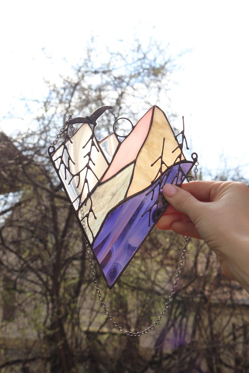 May include: A stained glass window hanging with a chain. The window depicts a mountain range with a pink, yellow, and blue color scheme. A small bird is flying above the mountains.