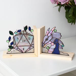 May include: Stained glass bookends featuring a D20 die and a dragon. The die is multi-colored with numbers, and the dragon is purple and blue. Both are set on wooden bases, perfect for a fantasy-themed bookshelf.