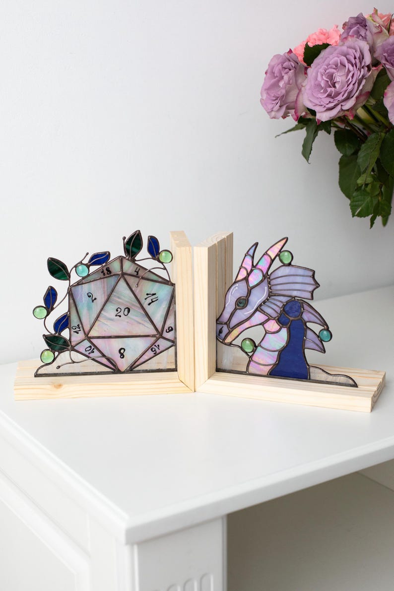May include: Stained glass bookends featuring a D20 die and a dragon. The die is multi-colored with numbers, and the dragon is purple and blue. Both are set on wooden bases, perfect for a fantasy-themed bookshelf.