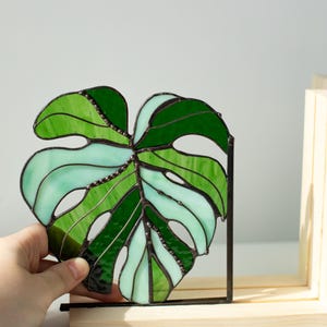 K&ouml;nnte beinhalten: Ein Buntglas-Monstera-Blatt in Gr&uuml;n- und Aquat&ouml;nen. Das Blatt wird in einem schwarzen Metallrahmen gehalten und in einem Holzst&auml;nder ausgestellt. Das Glas ist mit dunklen Linien segmentiert, wodurch ein detailliertes botanisches Design entsteht.