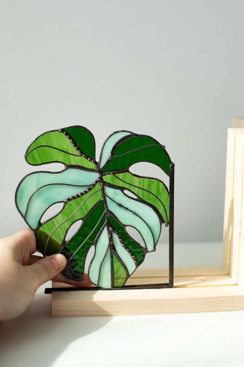 K&ouml;nnte beinhalten: Ein Buntglas-Monstera-Blatt in Gr&uuml;n- und Aquat&ouml;nen. Das Blatt wird in einem schwarzen Metallrahmen gehalten und in einem Holzst&auml;nder ausgestellt. Das Glas ist mit dunklen Linien segmentiert, wodurch ein detailliertes botanisches Design entsteht.
